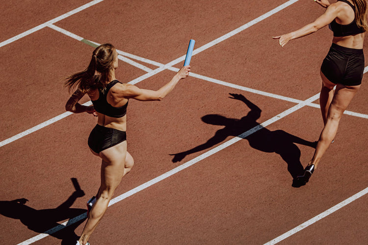 dwie kobiety biorą udział w biegu sztafetowym podają sobie pałeczkę